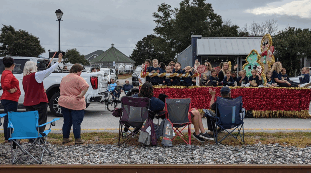Group of people gathered in a park watching a parade, with a gingerbread men float being pulled past and people waving to the crowd.